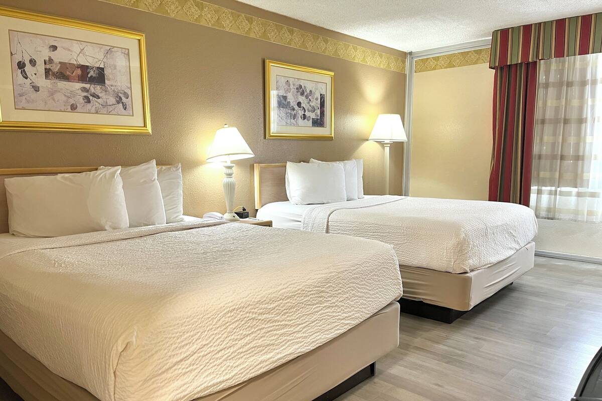 A hotel room with two beds, a large window with curtains, and two framed pictures on the wall.