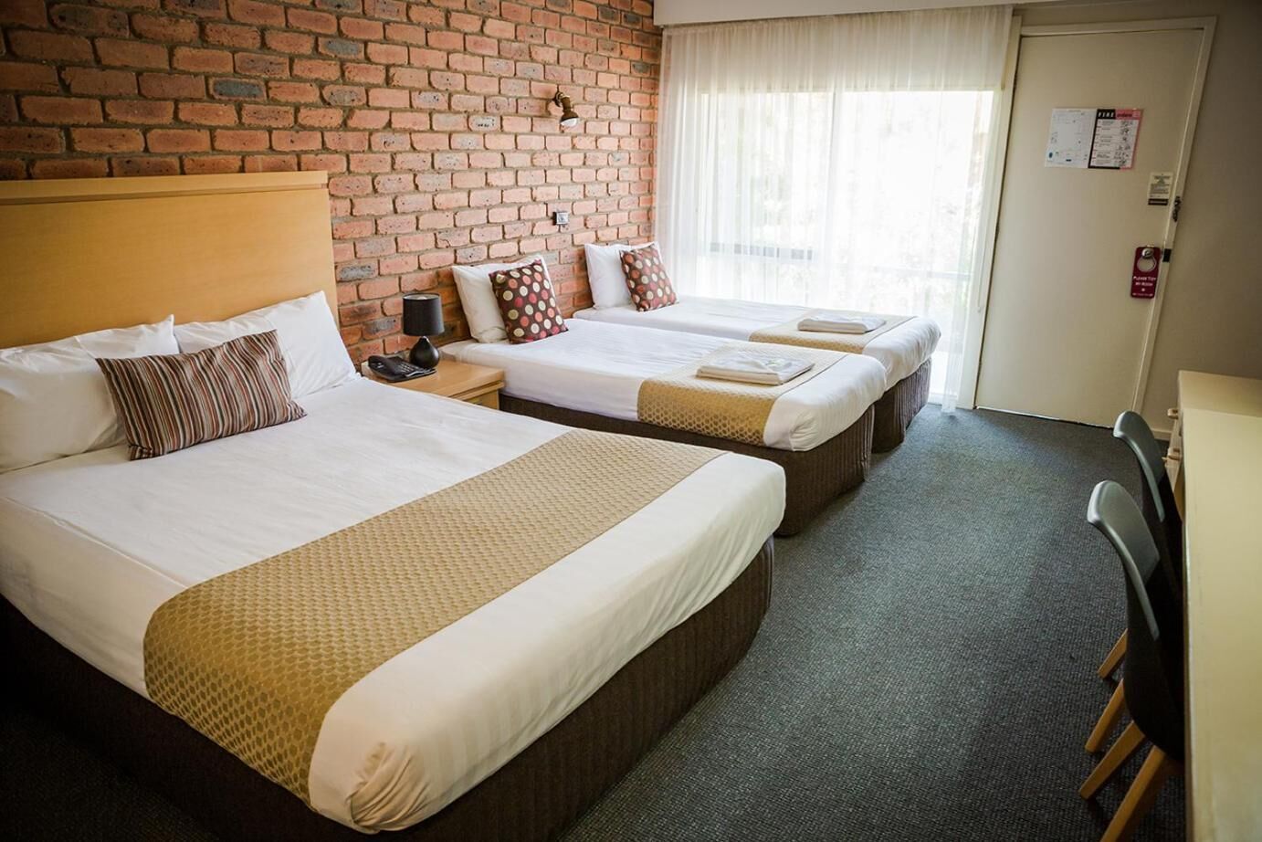 Habitación de hotel con dos camas, una pared de ladrillo, una ventana con cortinas, un escritorio y una silla.