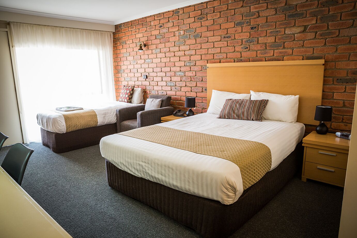 Une chambre d’hôtel avec deux lits, un mur en briques, une tête de lit en bois et un bureau avec une chaise.