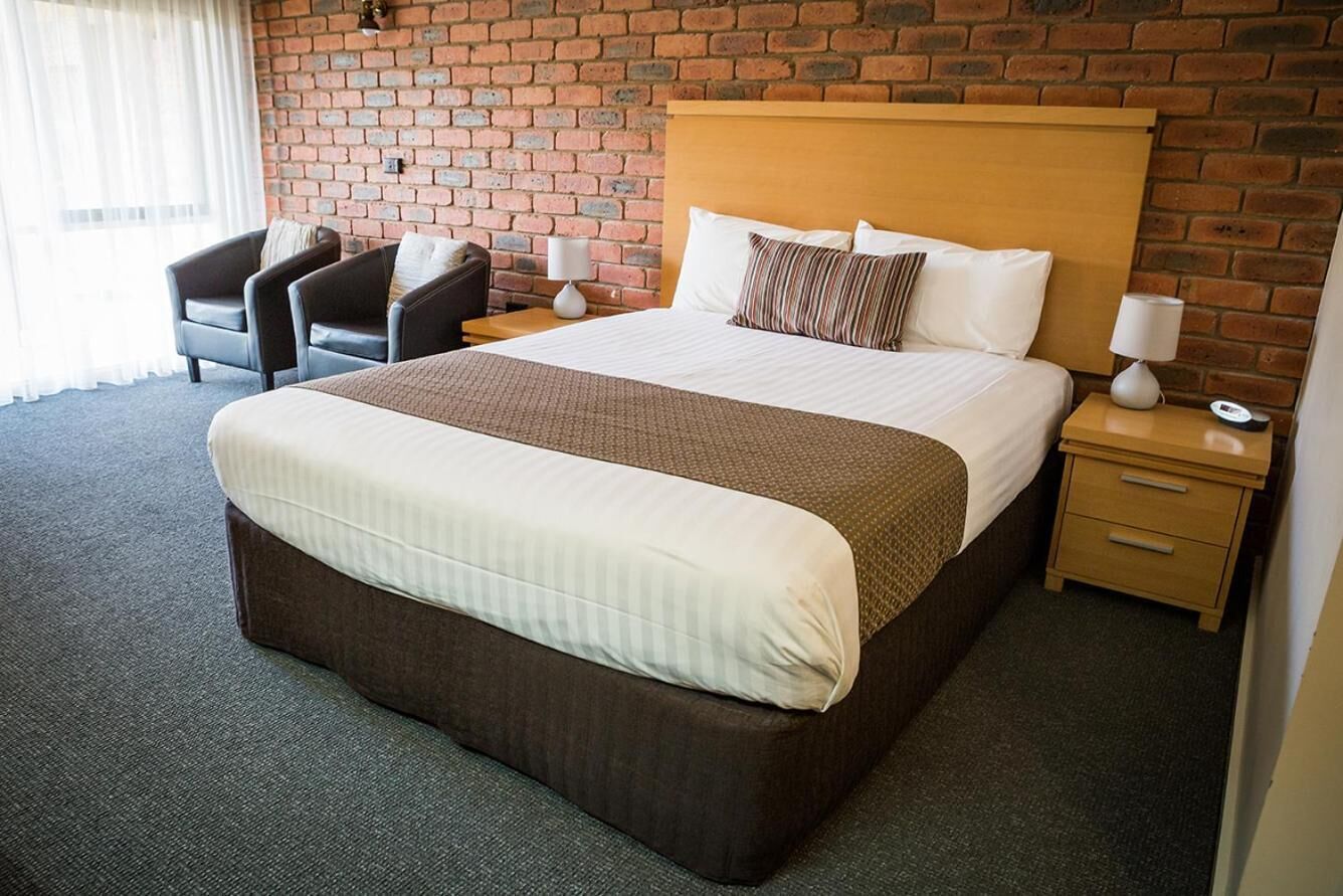 Une chambre d’hôtel avec un grand lit, une tête de lit en bois, un mur en briques, un bureau avec une lampe et une chaise.