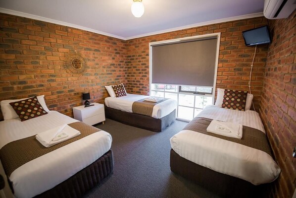 A hotel room with three beds, a brick wall, a large window, and a flat-screen TV. - Australian Settlers Motor Inn (Swan Hill)