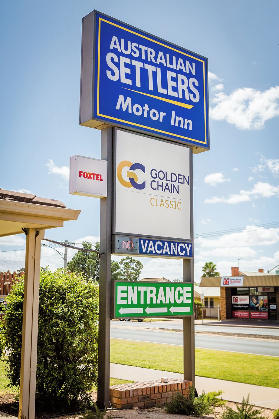Une enseigne indiquant l’emplacement de l’Australian Settlers Motor Inn, avec une annonce indiquant qu’il y a des chambres disponibles, des indications pour trouver l’entrée, et les noms d’entreprises situées à proximité, comme Foxtel et Golden Chain Classic.