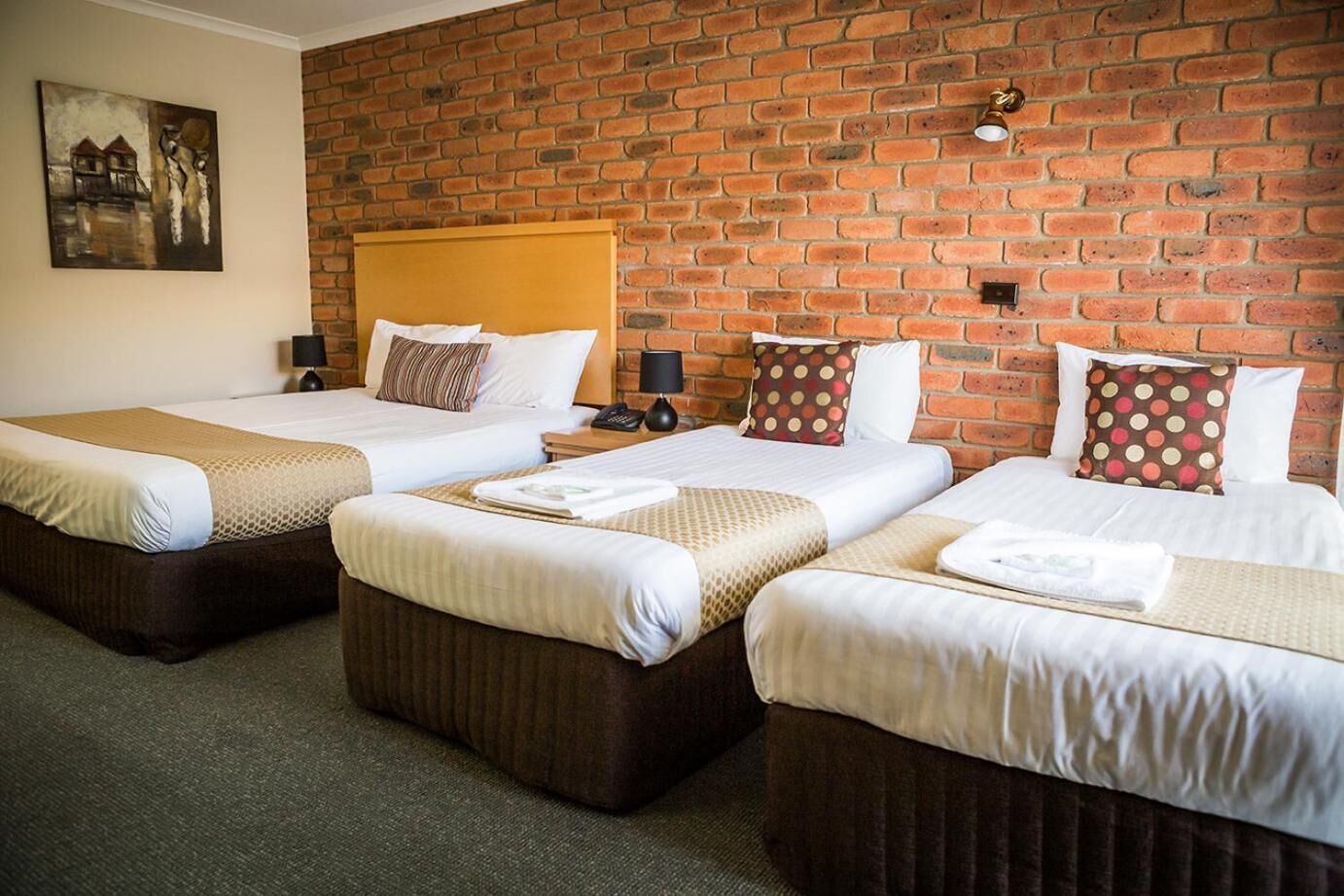 A hotel room with three beds, a brick wall, and a painting.