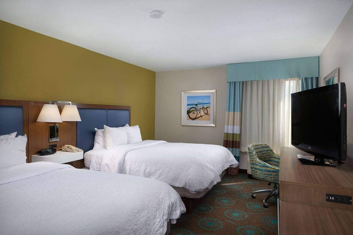 A hotel room with two beds, a TV, a chair, a painting, and a window with curtains.