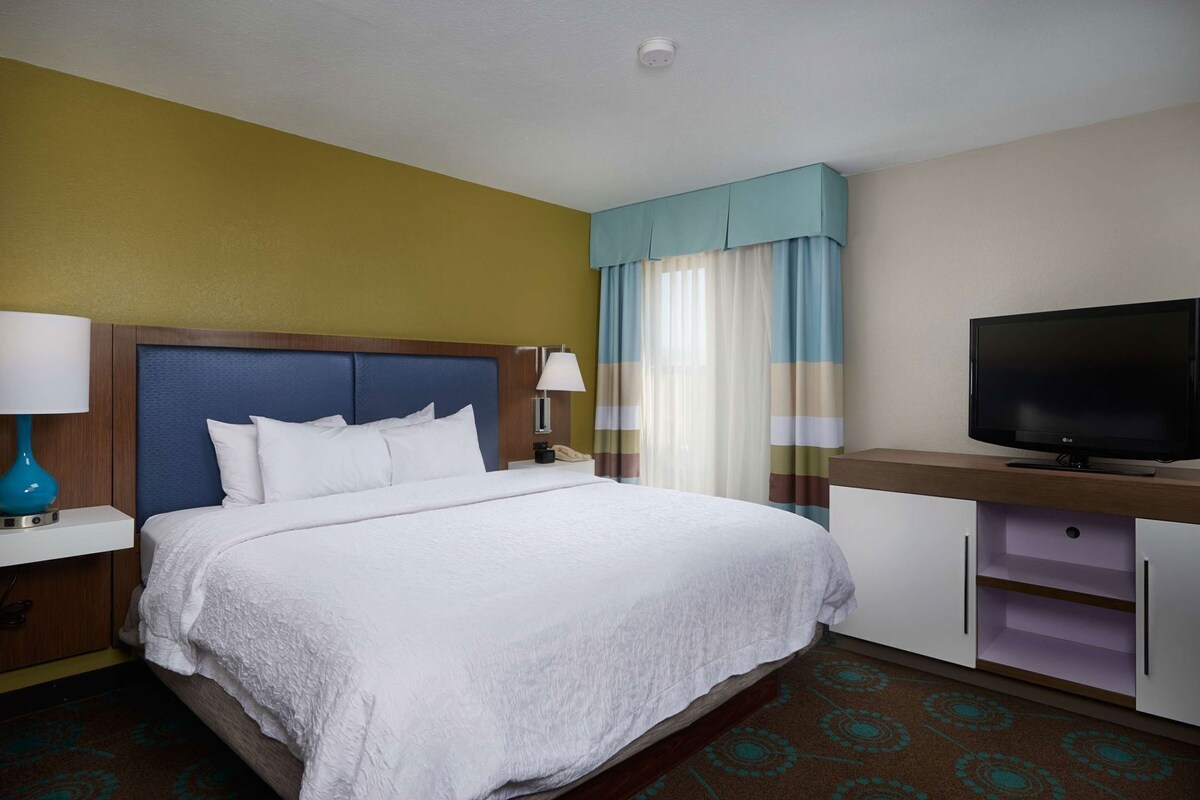 Une chambre d’hôtel avec un lit, une télévision, une table de chevet, une lampe et une fenêtre avec des rideaux.