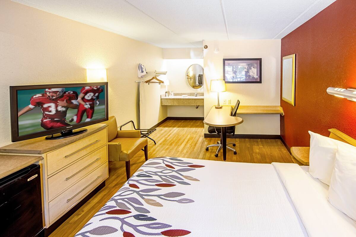 Ein Hotelzimmer mit einem Bett, einem Fernseher auf einer Kommode, einem Schreibtisch mit Stuhl, einem kleinen Tisch mit Lampe und einem Spiegel.