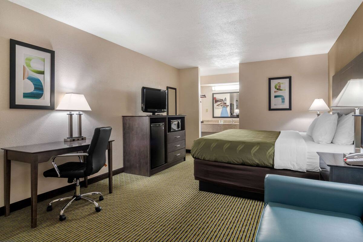 A hotel room with a bed, desk, chair, television, and a bathroom visible in the background.