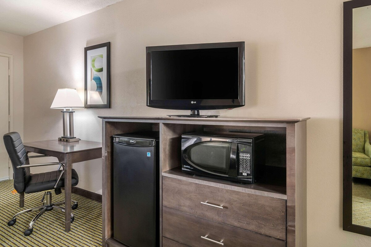 A hotel room with a flat-screen TV, a microwave, a mini-fridge, a desk with a chair, and a lamp.
