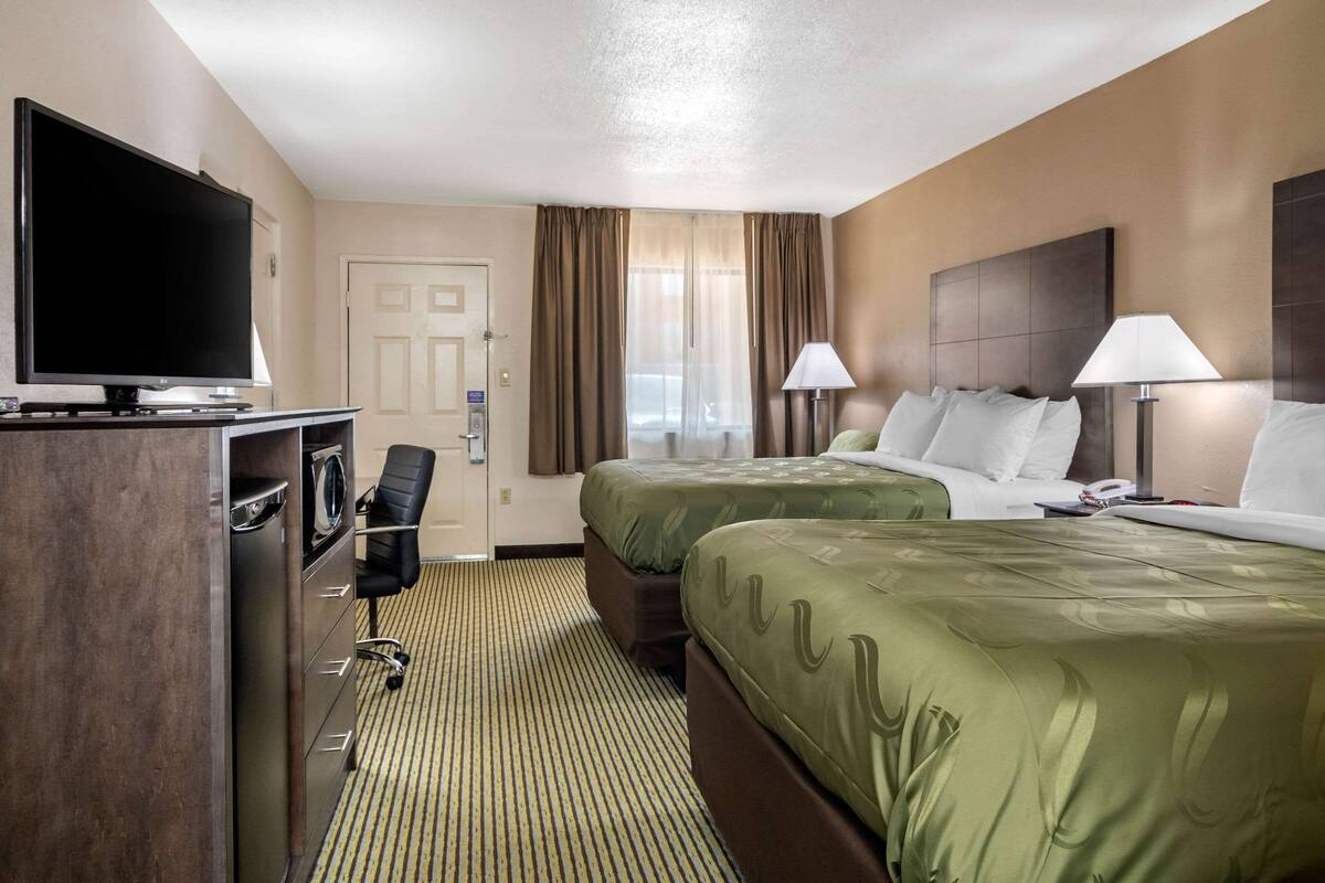A hotel room with two beds, a TV, a desk with a chair, and a lamp.