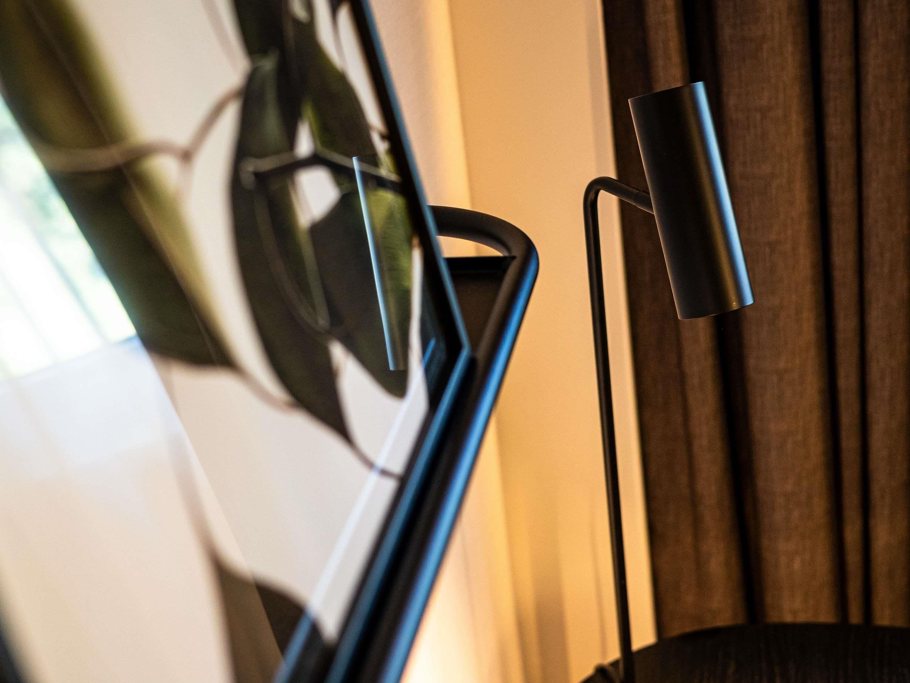 A modern lamp with a curved base and a cylindrical shade, positioned next to a window with a view of a plant.