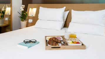 Uma cama bem arrumada com roupa de cama branca, uma bandeja de café da manhã com croissants, frutas e suco, e um livro na lateral.