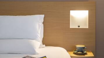 Een netjes opgemaakt bed met witte lakens, een klein nachtkastje met een kop koffie en een tijdschrift, en een wandlamp.