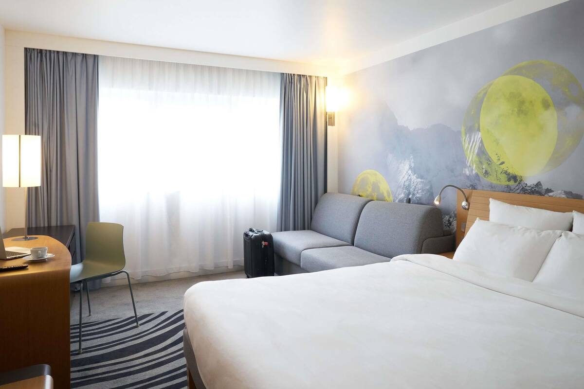 A hotel room with a bed, a sofa, a desk with a laptop, a chair, and a wall mural of a moon.