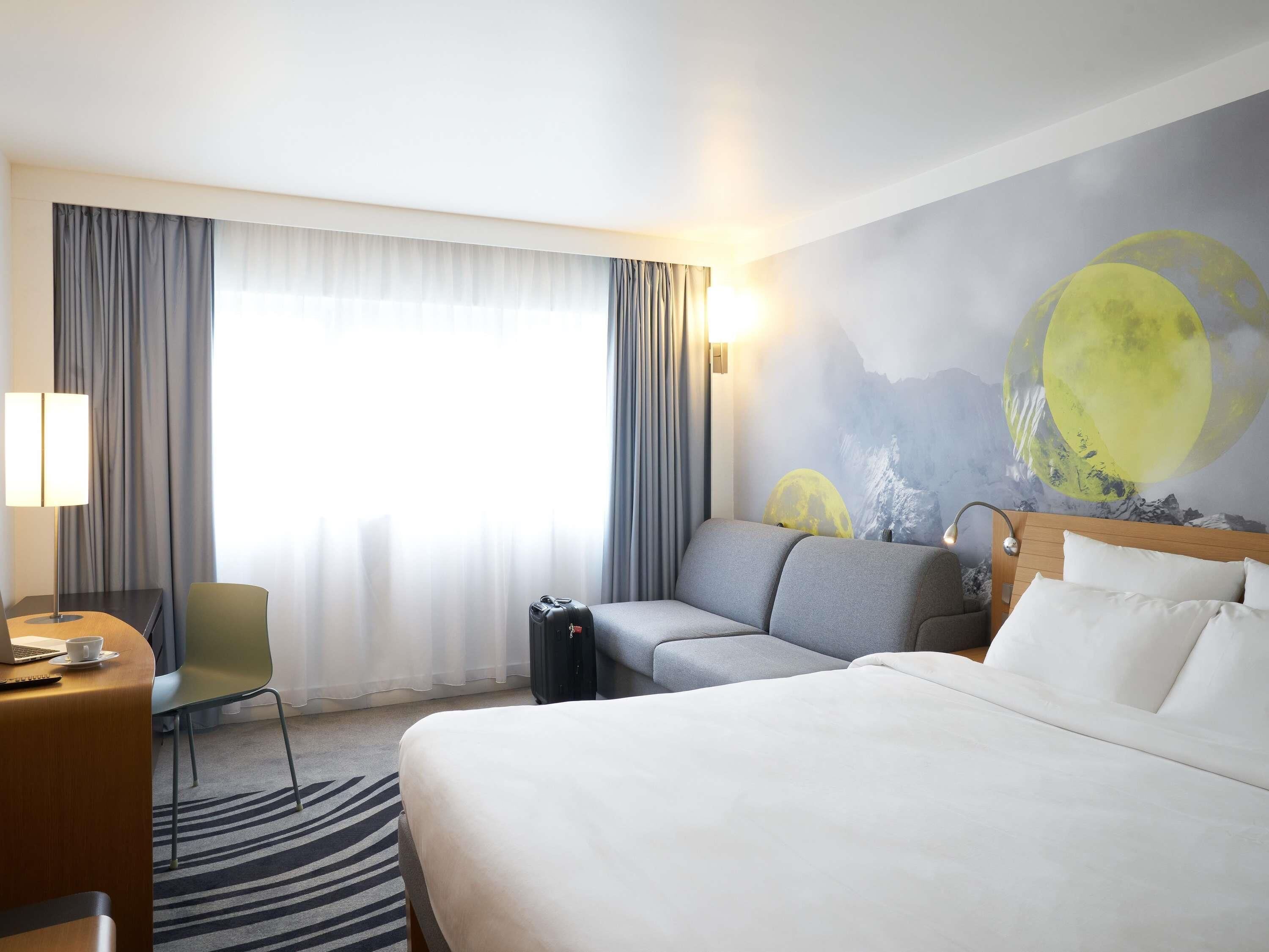 A hotel room with a bed, a sofa, a desk with a laptop, a chair, and a wall mural of a moon.