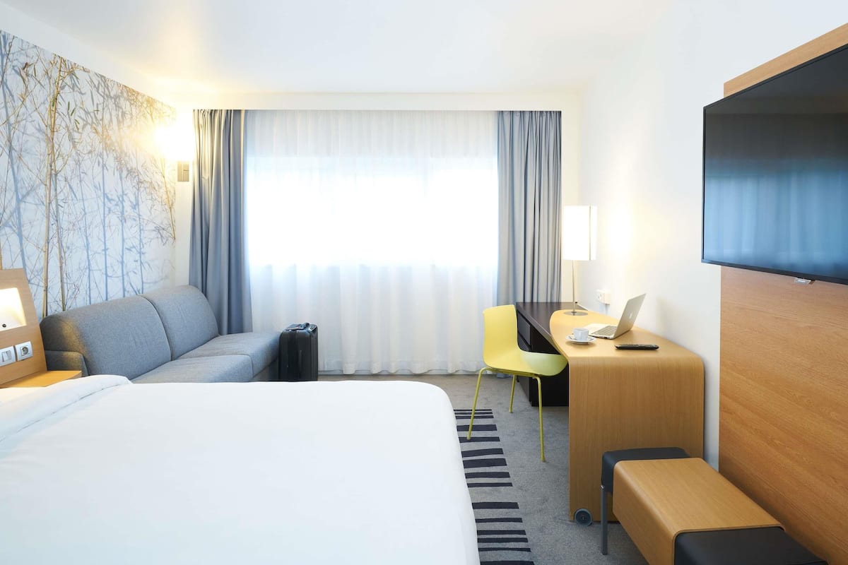 A hotel room with a bed, a desk with a laptop, a chair, a TV, and a wall with a nature-themed design.