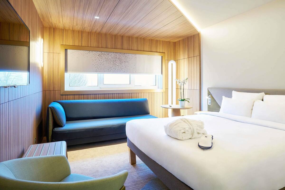 A modern hotel room with a wooden ceiling, a bed with white linens, a blue sofa, a small table with a plant, and a window with blinds.