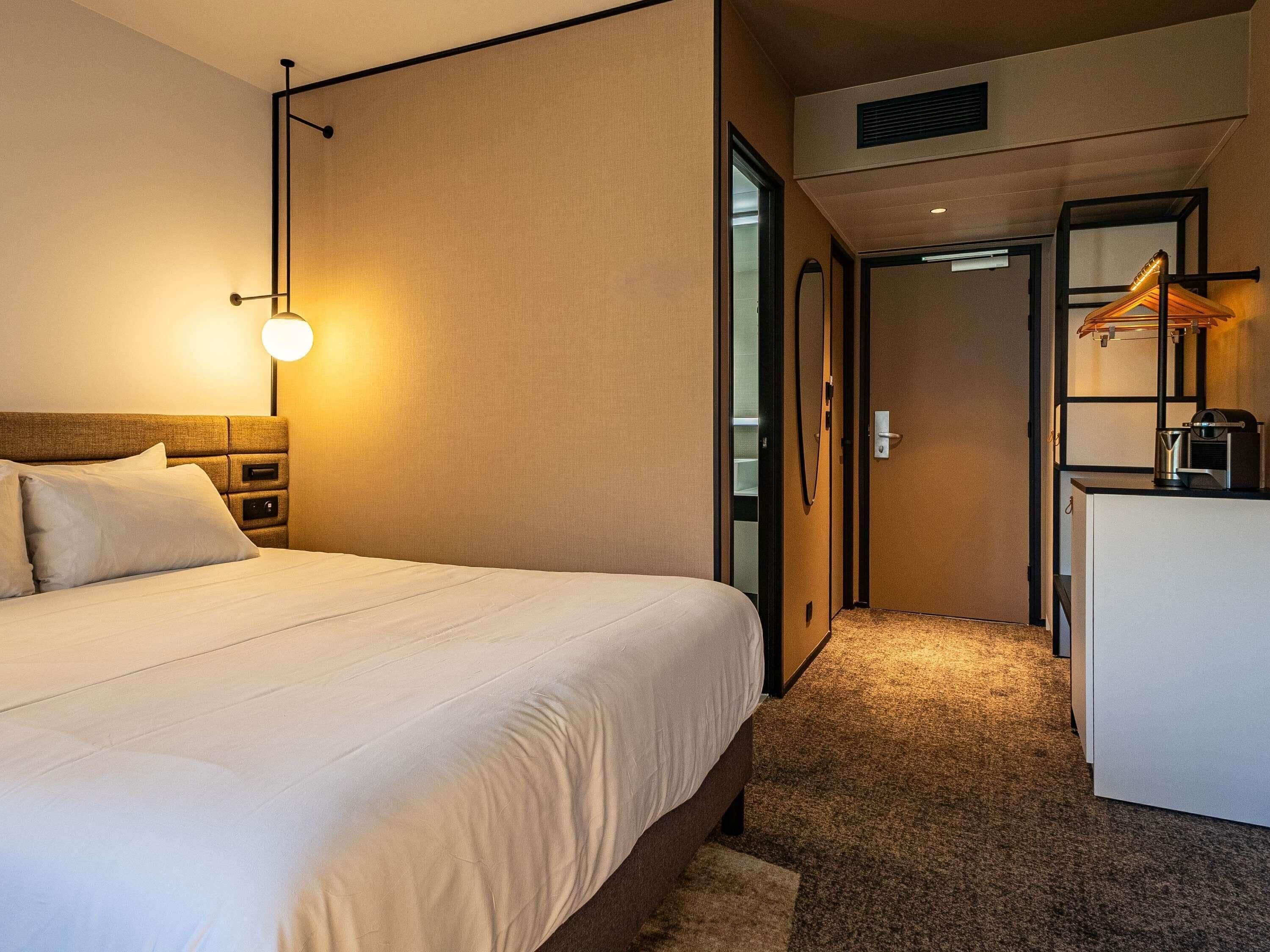 A hotel room with a bed, bedside table, lamp, and a desk with a mirror and a kettle.