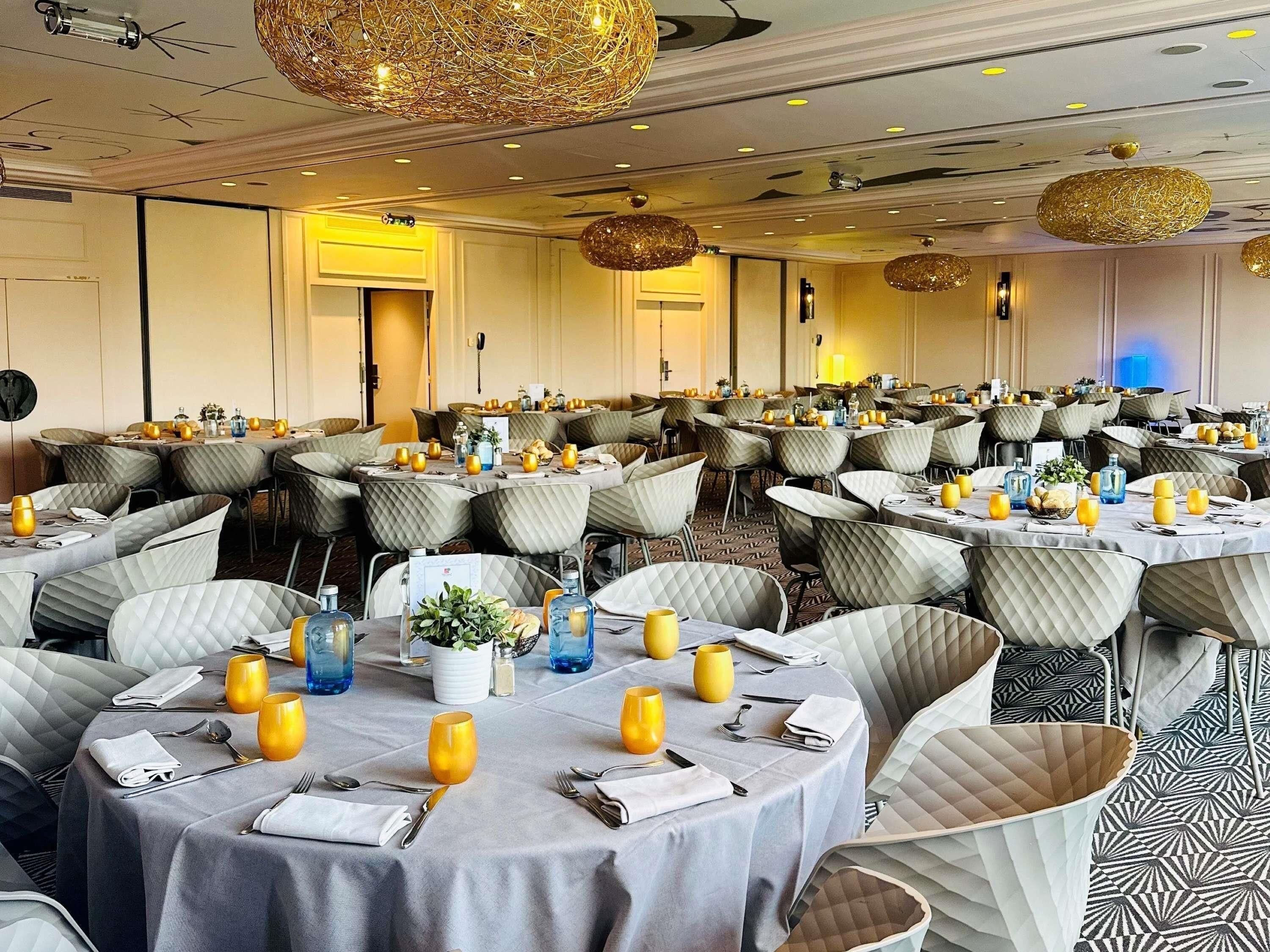 A banquet hall with round tables, cushioned chairs, and hanging spherical light fixtures.