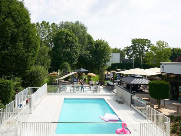 A swimming pool with a pink flamingo float, surrounded by outdoor seating and umbrellas, with trees and a building in the background. - Novotel Marne Vallee Collegien (Collegien)