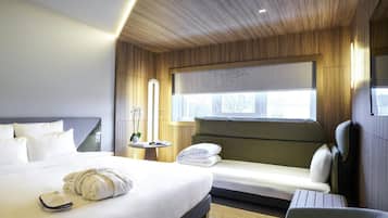 A hotel room with a large bed, a smaller sofa, a chair, a bedside table with a lamp, and a window with a view.
