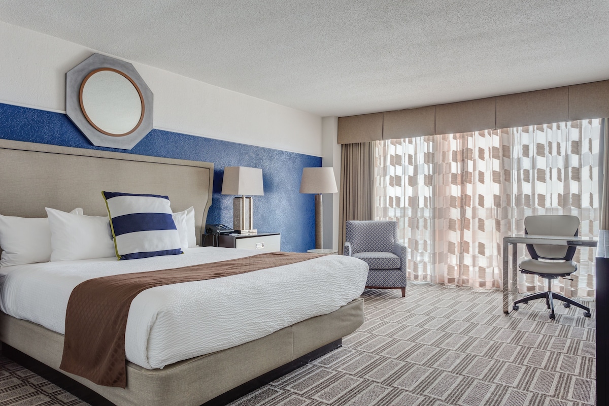 A hotel room with a large bed, two bedside tables, a desk, a chair, and a window with patterned curtains.
