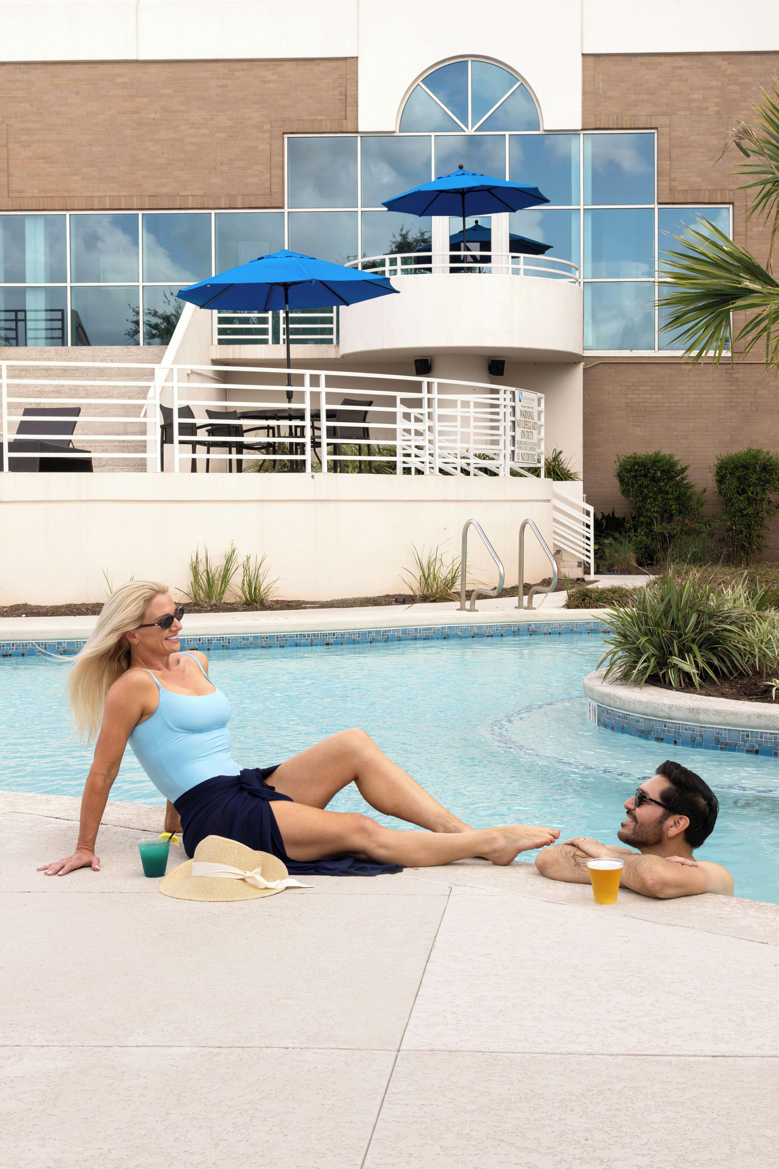 A swimming pool with a modern building in the background, a lounge area with chairs, and a palm plant.