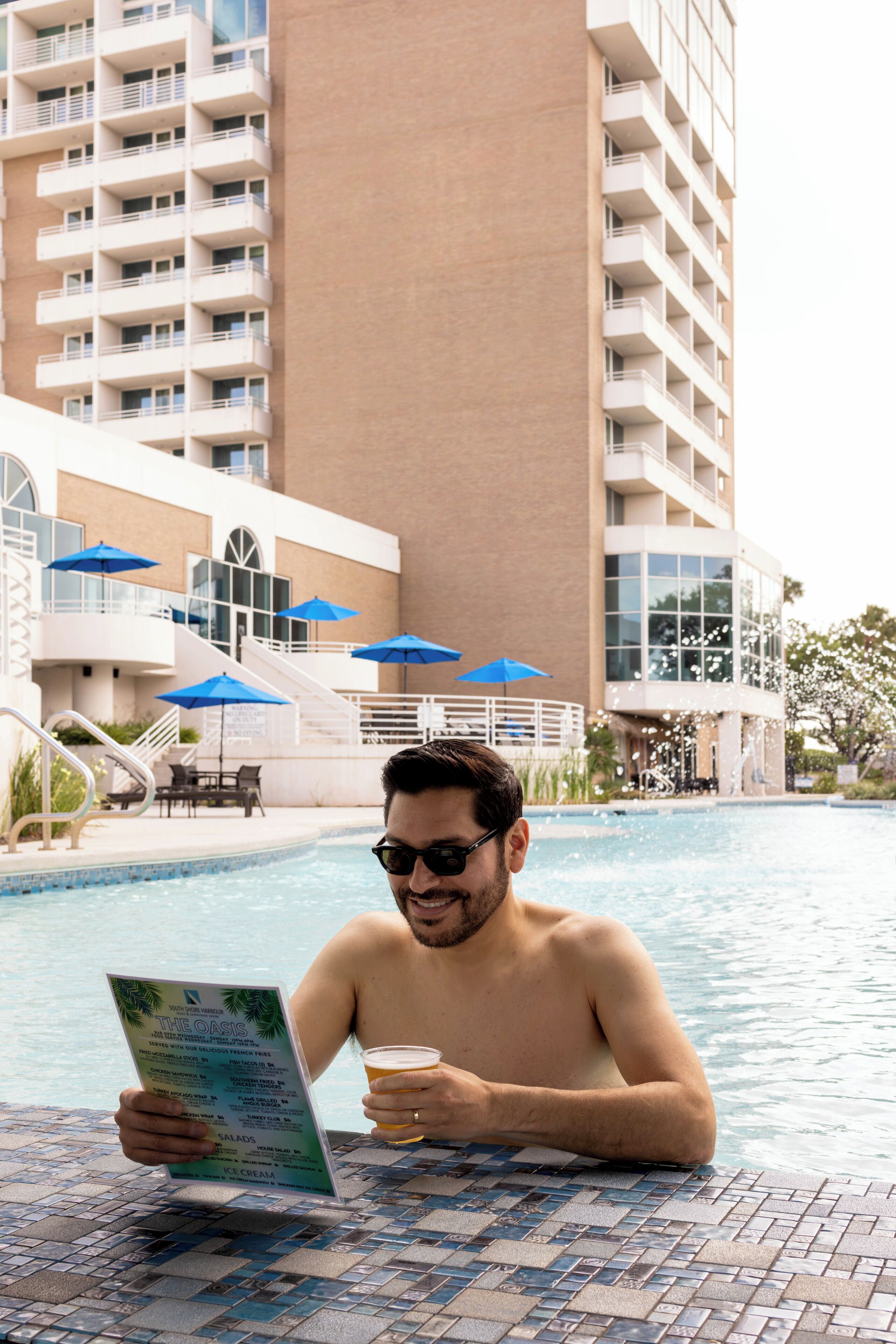 A man in a swimming pool reading a menu, with a tall building and blue umbrellas in the background.