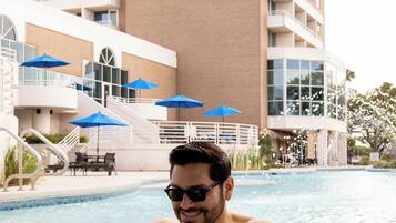 A man in a swimming pool reading a menu, with a tall building and blue umbrellas in the background.