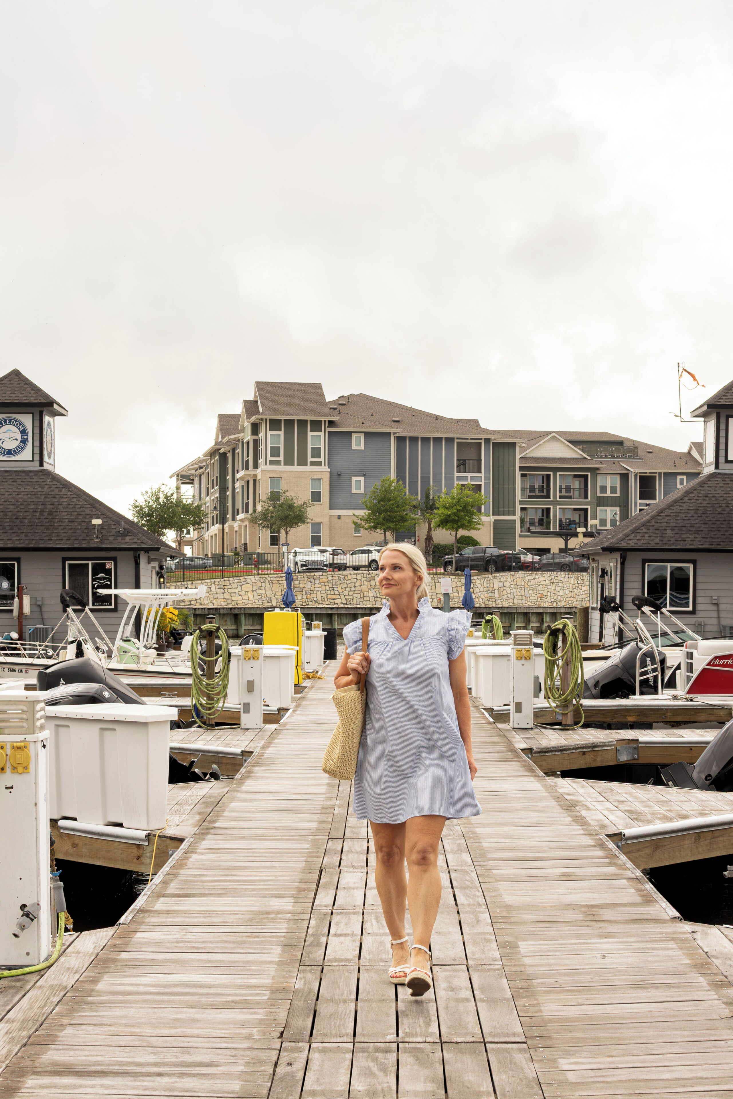 A wooden pier leading to a marina with boats docked, a clock tower, and residential buildings in the background.