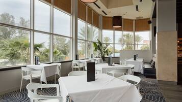 Un ristorante con tavoli e sedie bianche, ampie finestre che si affacciano su palme e lampade pendenti.