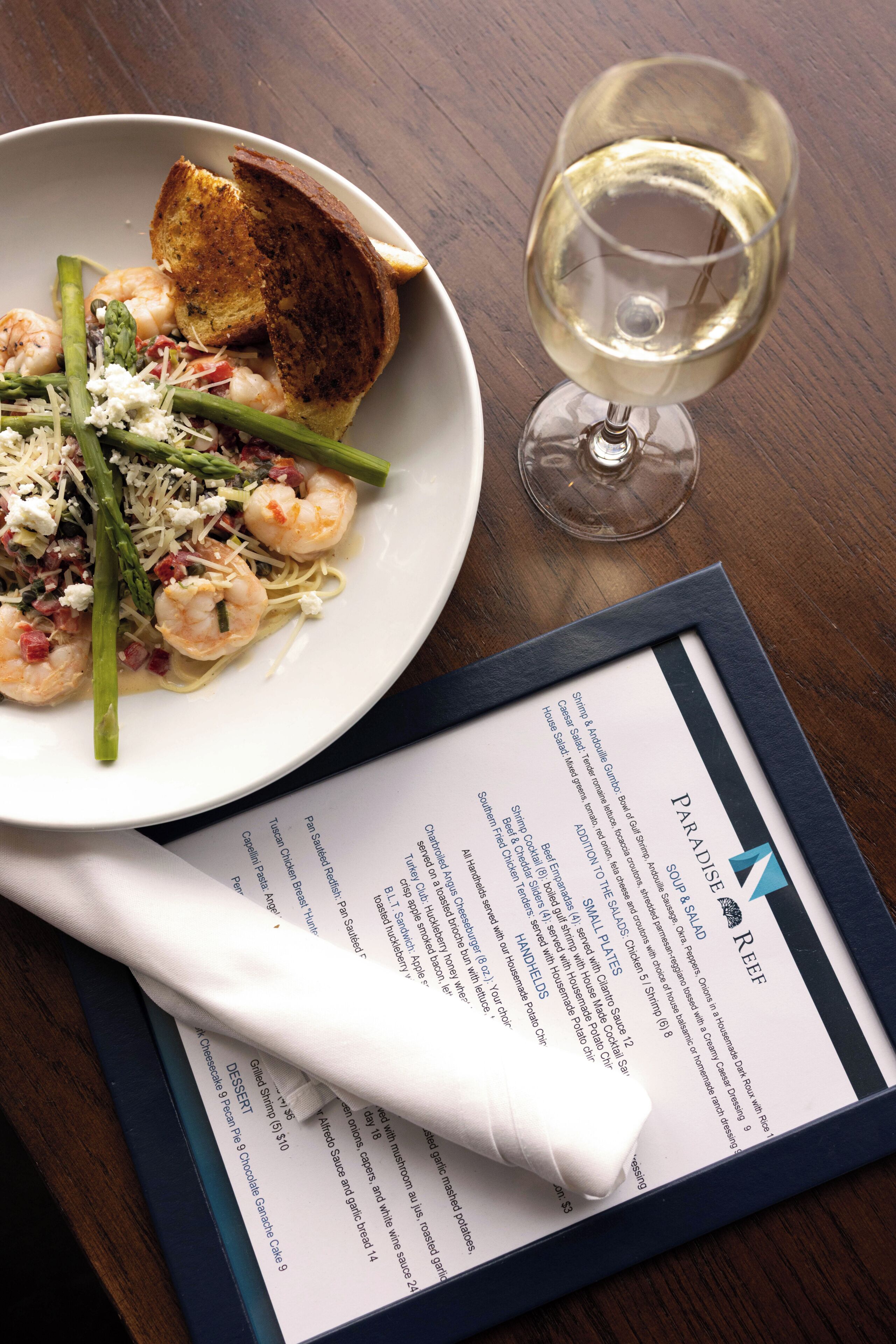 Une assiette de nourriture comprenant des crevettes, des asperges et une tranche de pain grillé, un verre de vin blanc et un menu posé sur une table en bois.