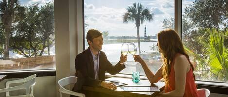Un homme et une femme trinquent avec des verres de vin, assis à une table d’où l’on peut admirer un lac et des palmiers.