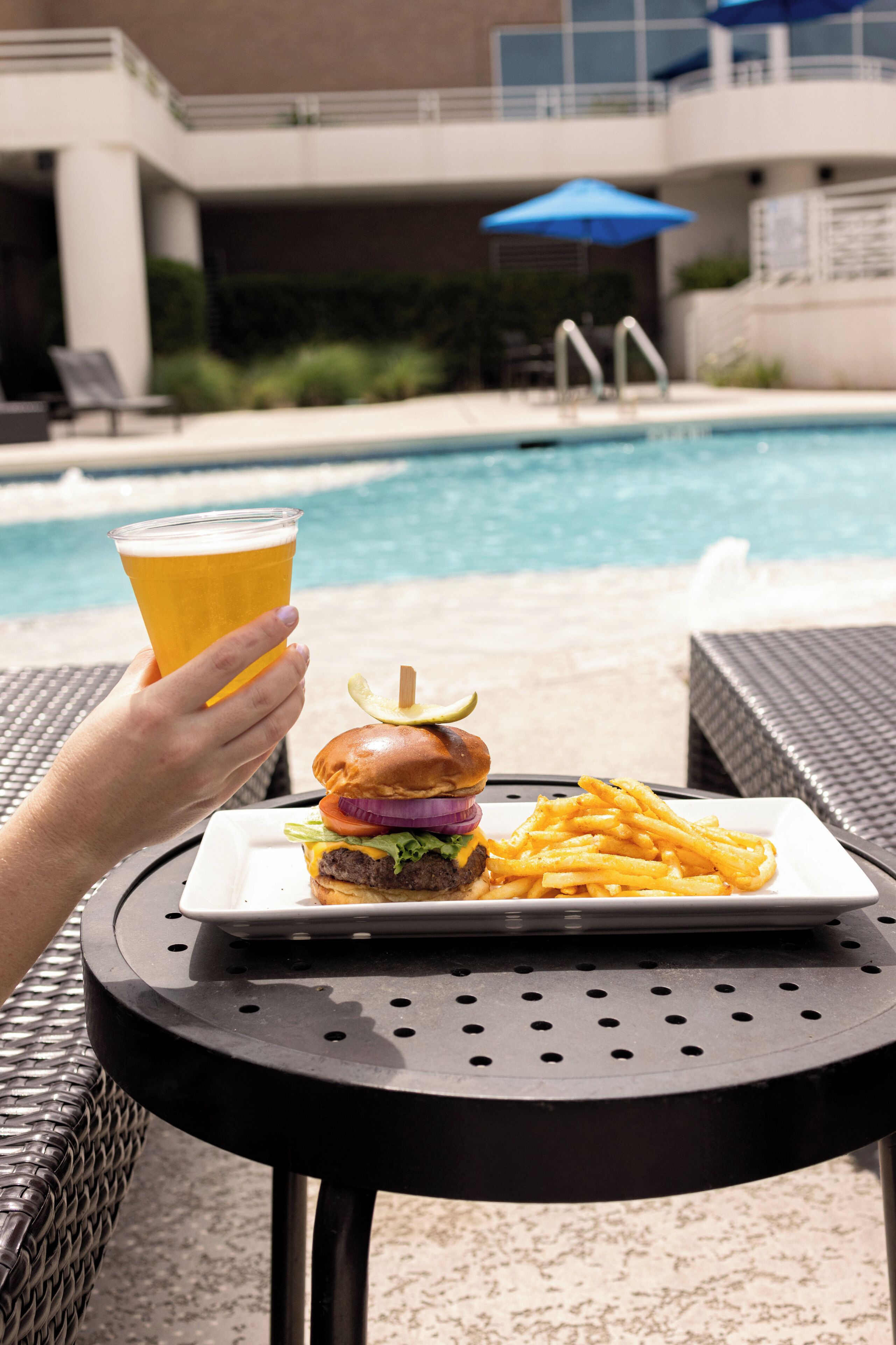 A dining table with a burger and fries, a glass of beer, and a pool in the background.