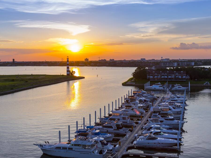 Un puerto deportivo con barcos amarrados, un atardecer de fondo y el perfil de una ciudad al otro lado del agua.