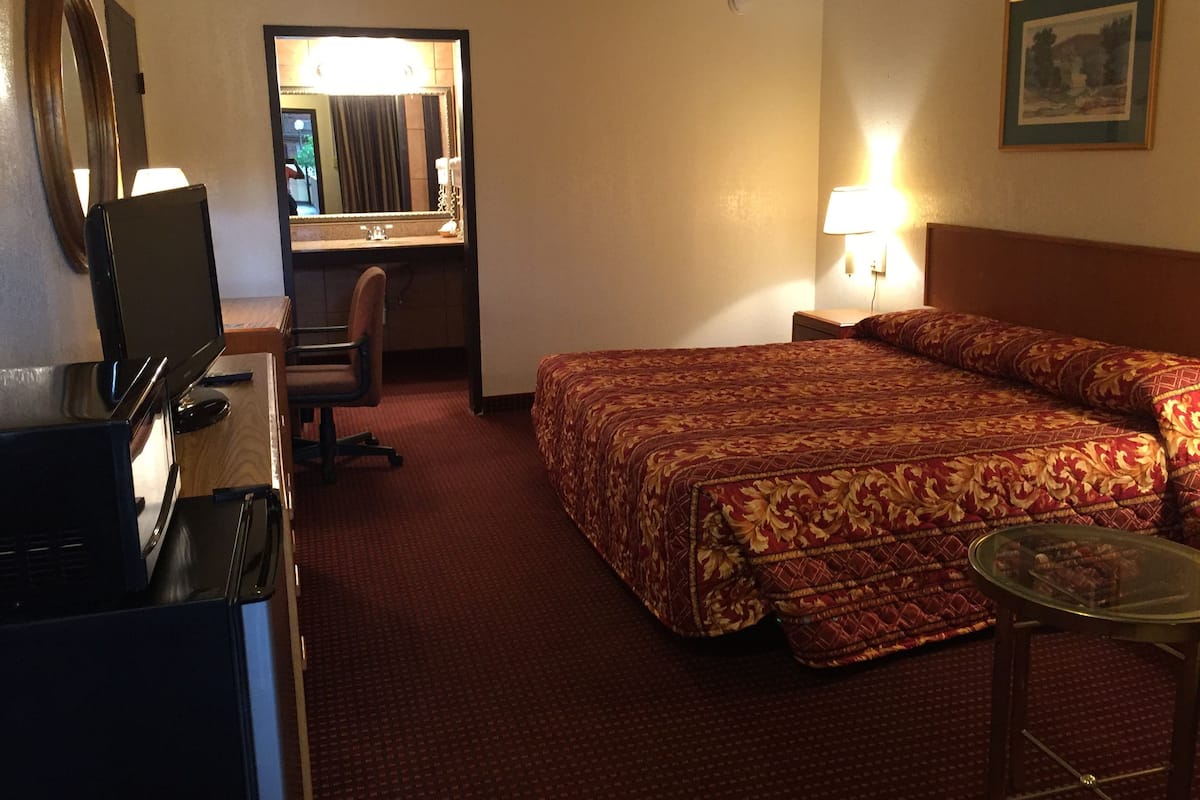 Quarto de hotel com uma cama, uma escrivaninha com cadeira, televisão, espelho e uma mesinha.