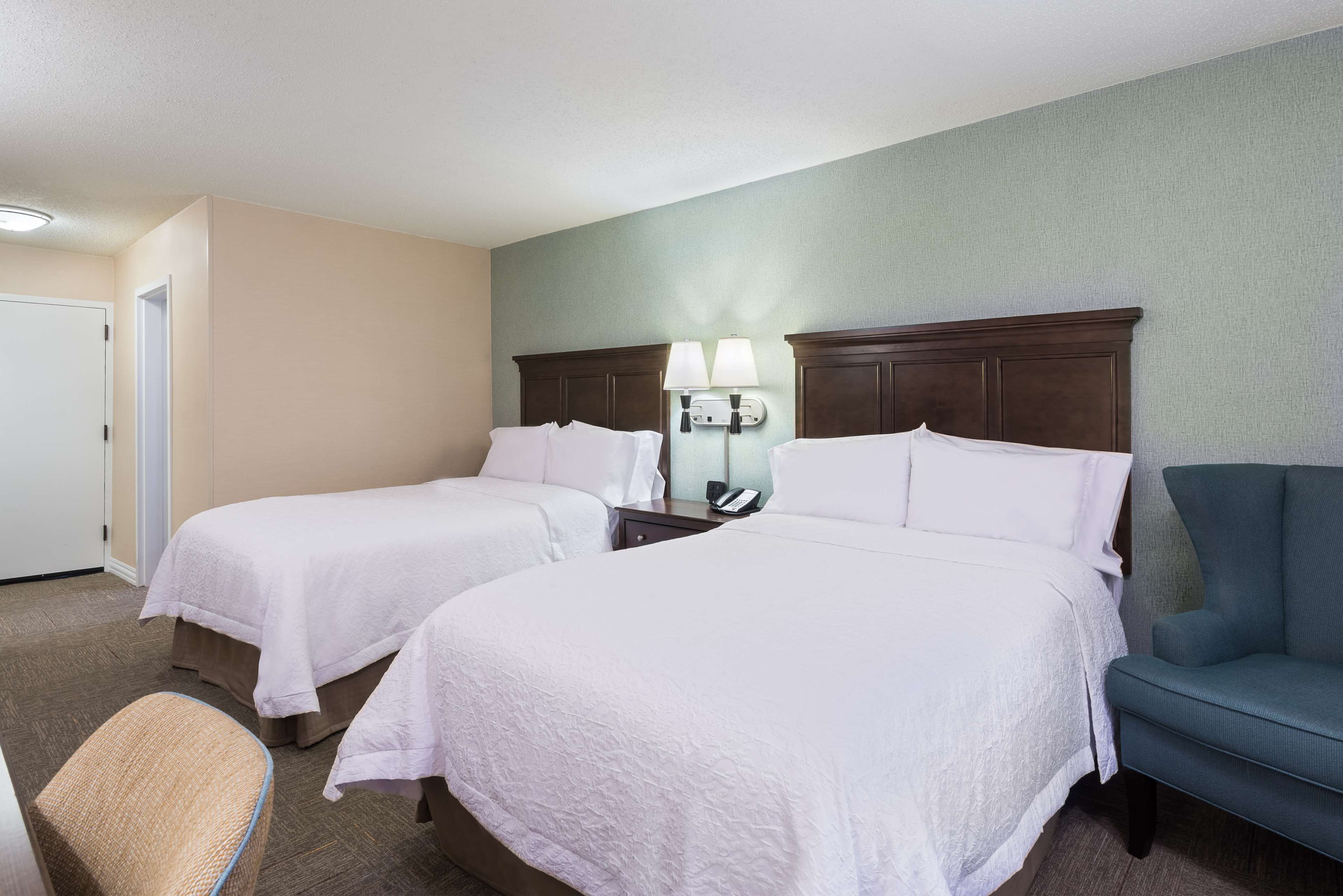 Una habitación de hotel con dos camas, una silla, una mesita de noche con lámpara y un teléfono.