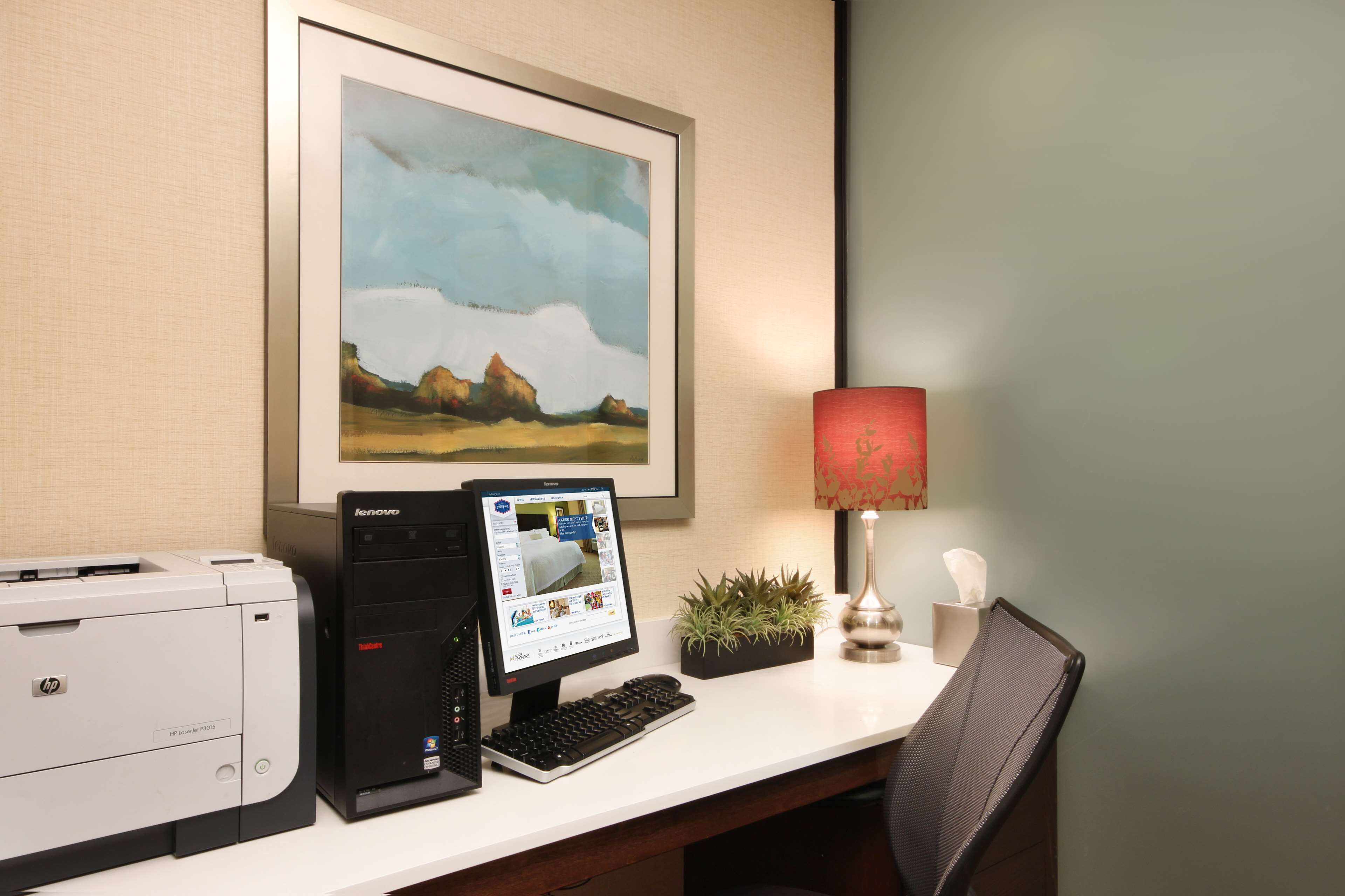 Een hotel receptie met een computer, een printer, een schilderij van een landschap, een rode lamp en een kleine plant.