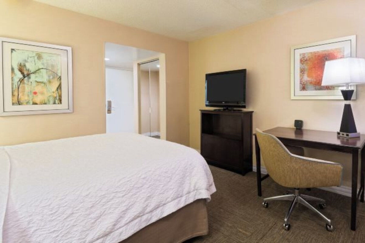 A hotel room with a large bed, a desk with a chair, a television, and two framed artworks on the walls.