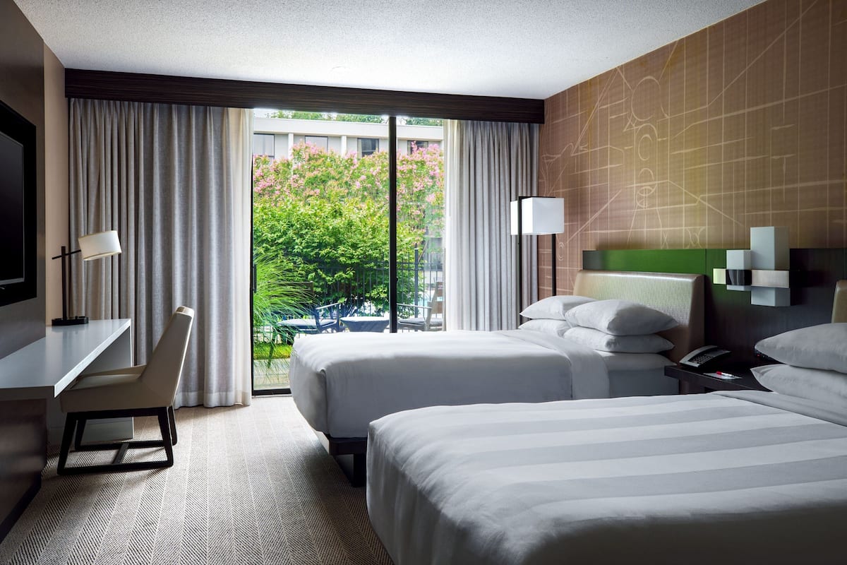 A hotel room with two beds, a desk, and a chair. There is a large window with curtains, and a view of greenery outside.