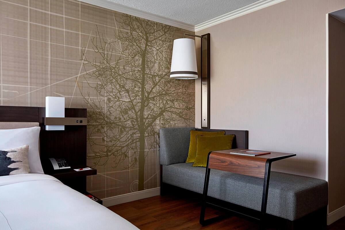 A modern hotel room with a bed, a gray sofa with a yellow pillow, a small wooden table, and a wall with a tree mural.
