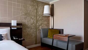 Ein modernes Hotelzimmer mit einem Bett, einem grauen Sofa mit gelbem Kissen, einem kleinen Holztisch und einer Wand mit einer Baummalerei.