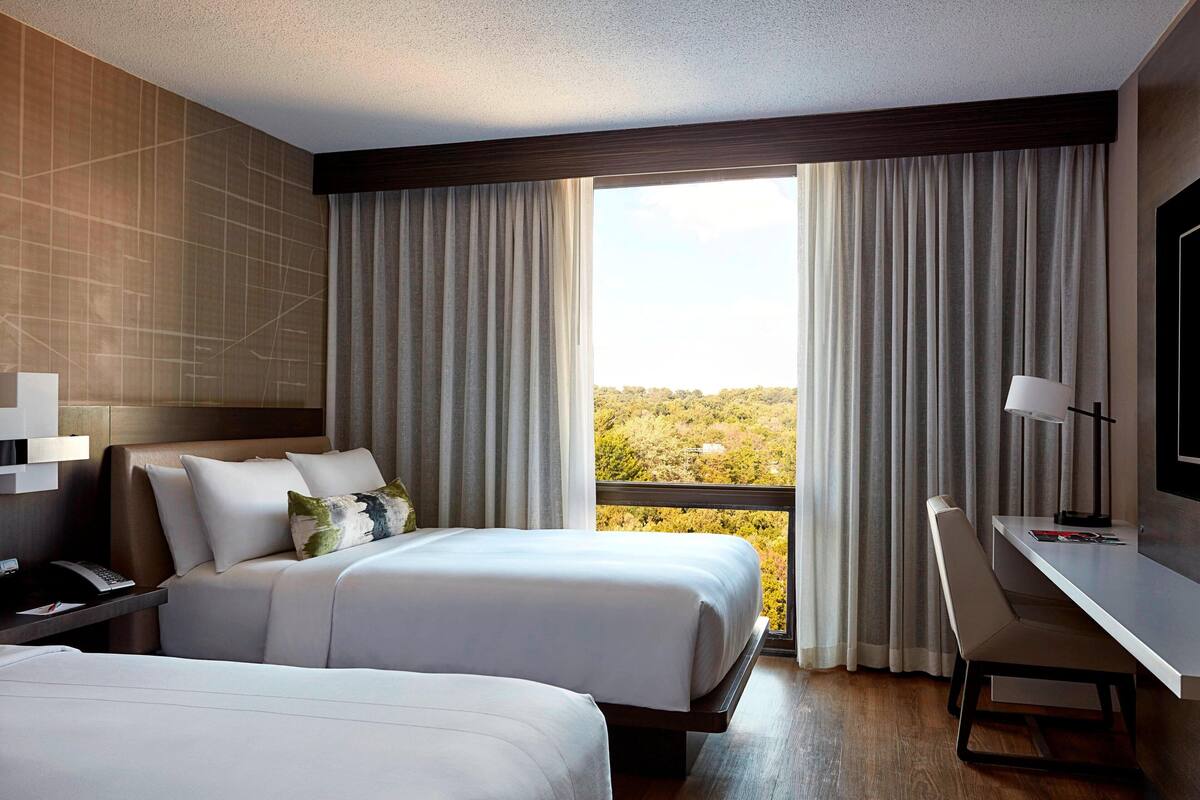 A hotel room with two beds, a desk, a chair, a TV, and a large window with curtains.