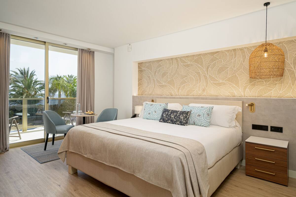 A modern hotel room with a large bed, a small desk, and a balcony with a view of palm trees.