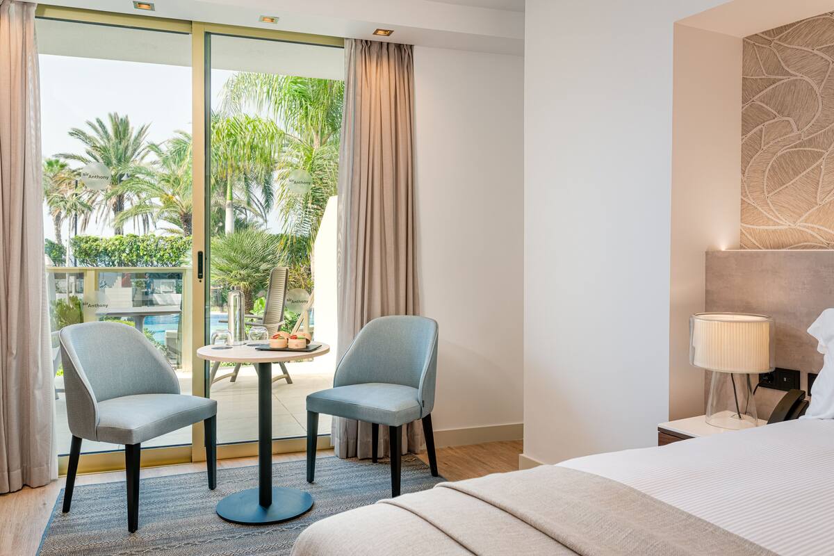 A hotel room with a bed, two chairs, a small table, a lamp, and a view of palm trees through a sliding door.