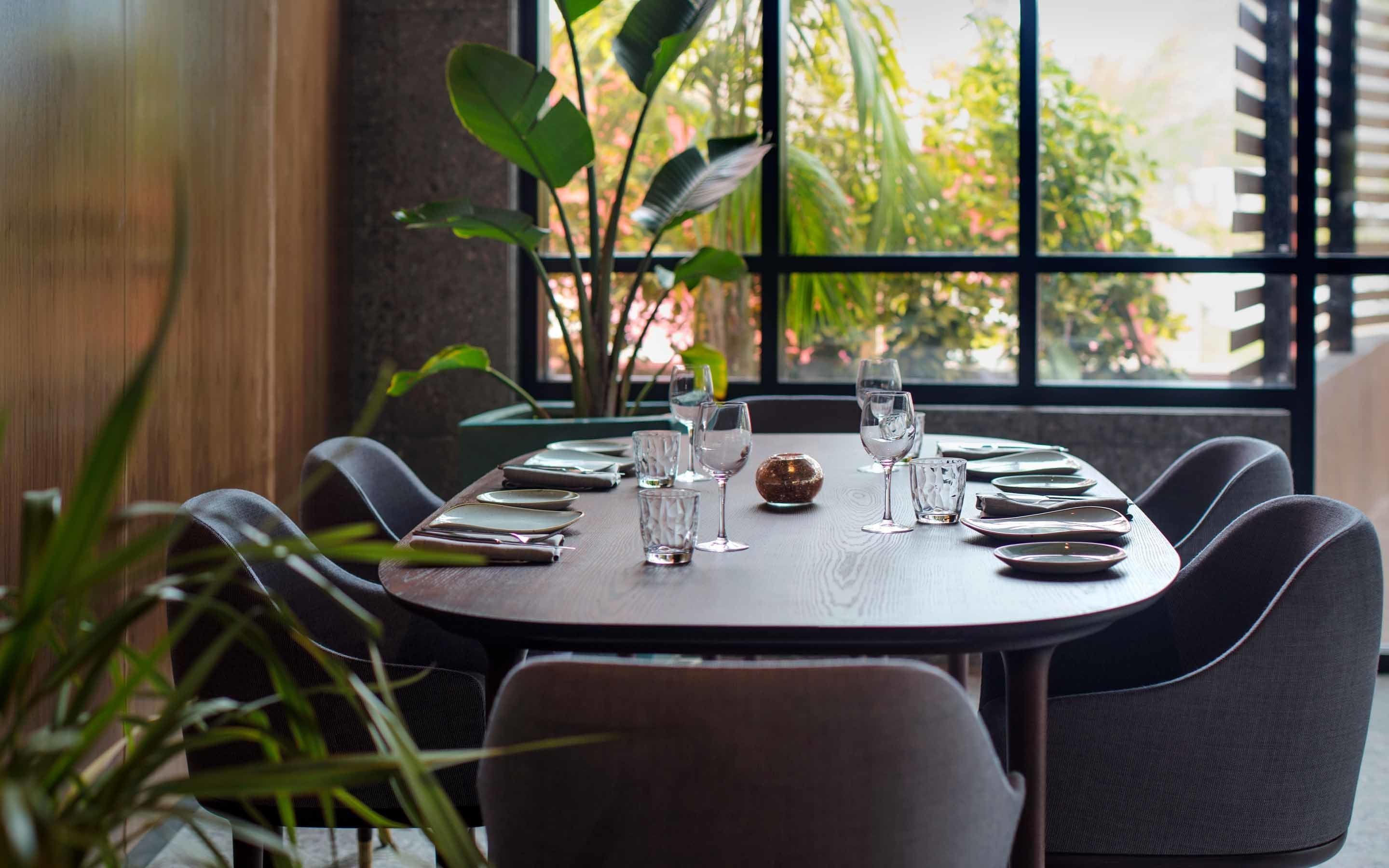 Een eettafel voor zes personen, gedekt met borden, glazen en bestek, staat bij een raam met uitzicht op groen.