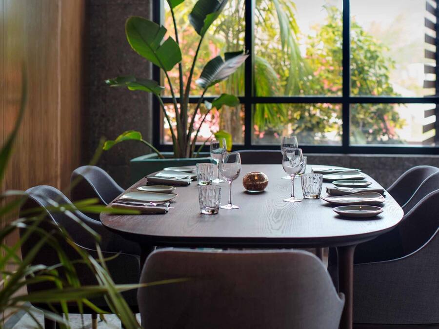 A dining table set for six with plates, glasses, and cutlery, positioned near a window with a view of greenery.