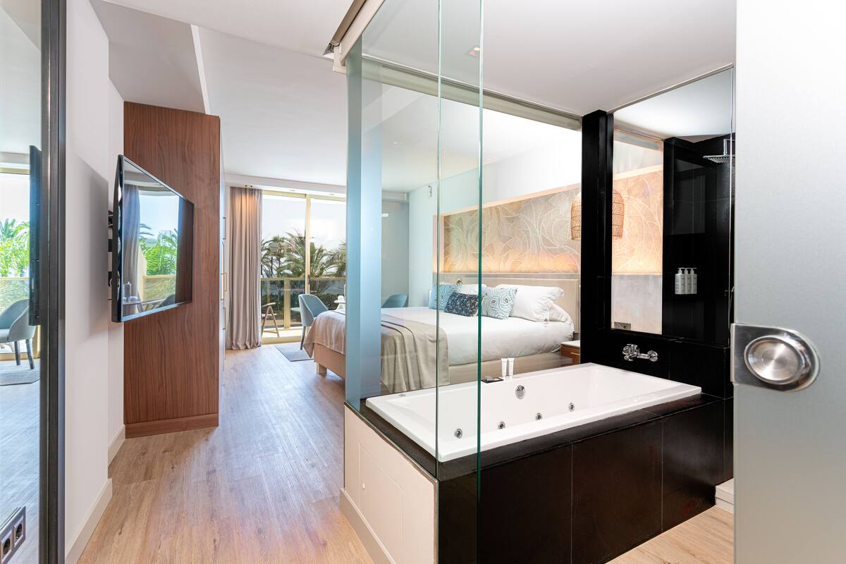 A modern hotel room with a large bed, a glass-enclosed shower, and a flat-screen TV mounted on the wall.