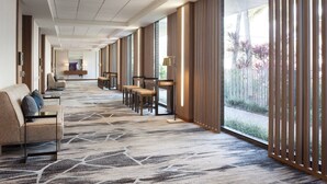 Ein Hotelkorridor mit einem gemusterten Teppich, Sitzbereichen und großen Fenstern mit Blick auf Palmen.