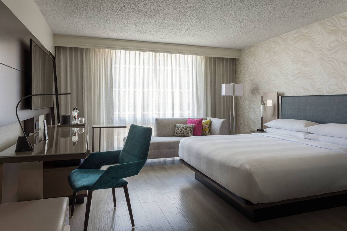 Habitación de hotel con cama, escritorio, silla, televisor y ventana con cortinas.