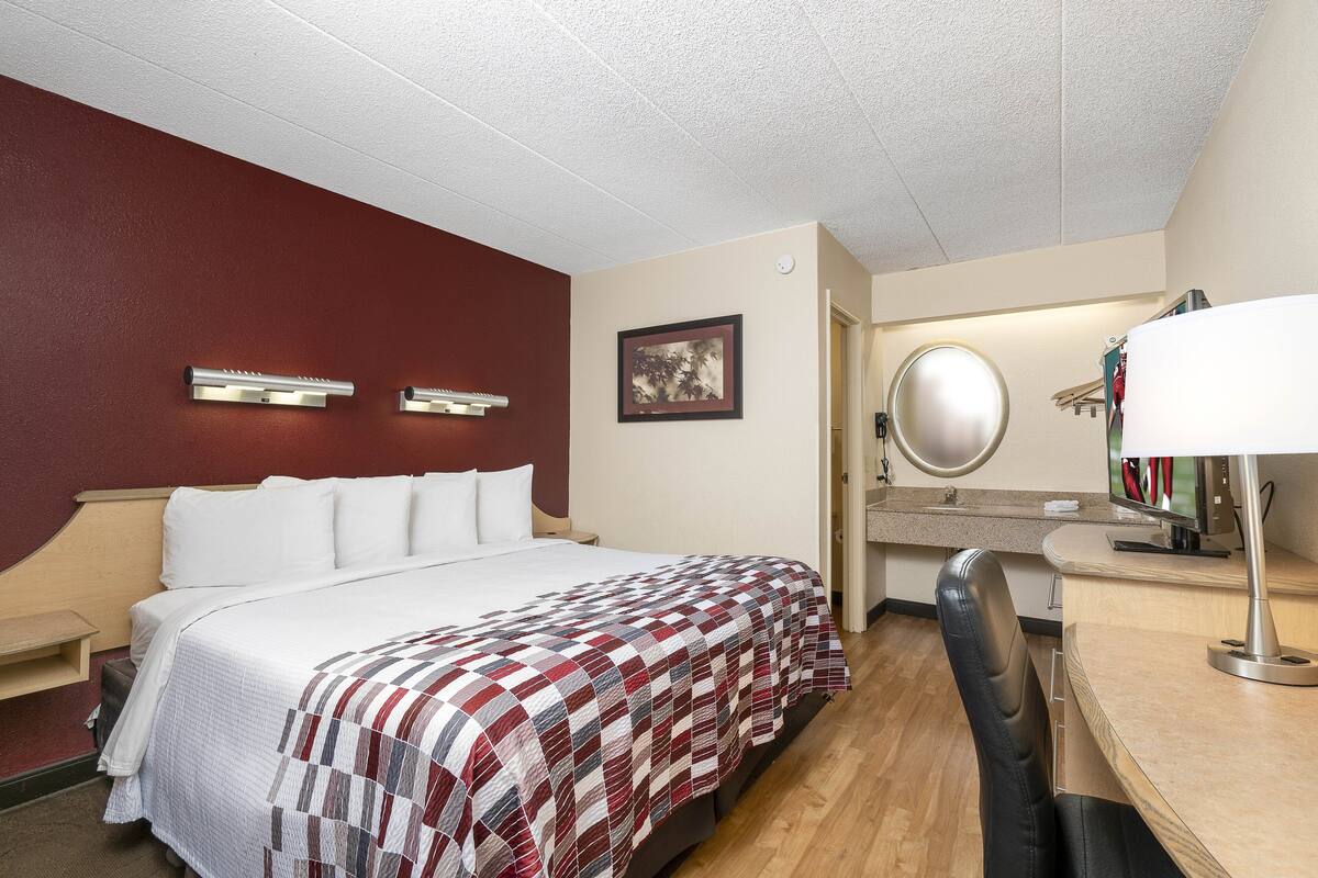 Una habitación de hotel con cama, escritorio, silla, espejo y un cuadro en la pared.